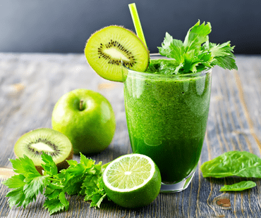 green fruit shake in a glass, next to lies an apple, cut into half a lime and kiwi; shake is decorated with mint, kiwi slice and straw